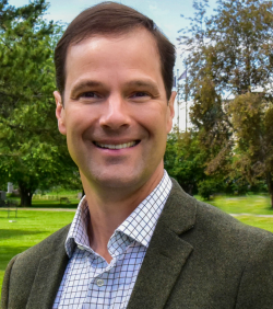 Kevin Pendergast, wearing a blue shirt and brown jacket, smiles in front of a green field.
